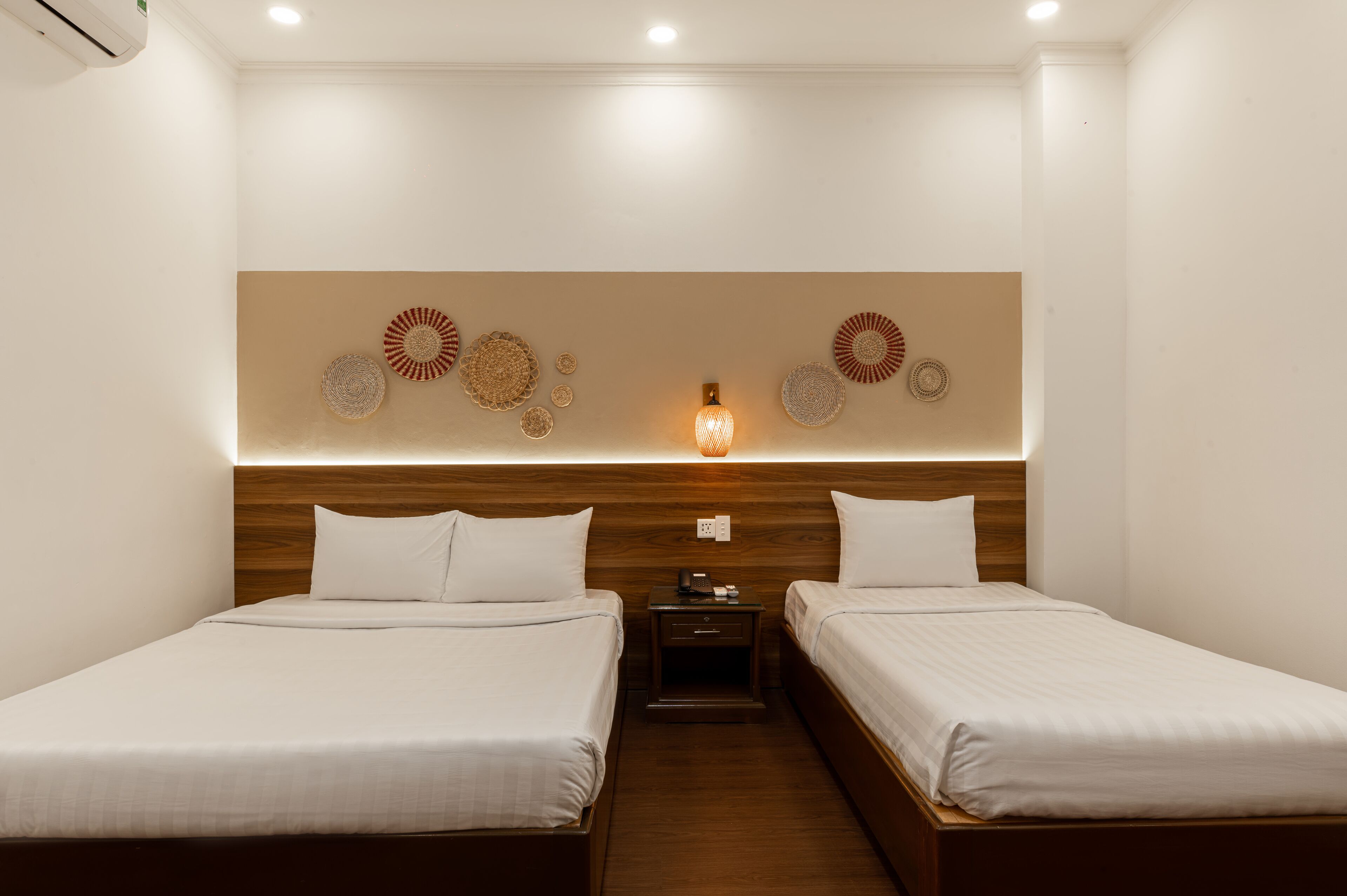 A hotel room with two beds, a wooden headboard, wall-mounted decorative plates, a nightstand, and a wall-mounted light fixture.