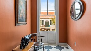 A hallway with a checkered floor, a wooden chair, a framed painting, a round mirror, and a view of a building through a door.