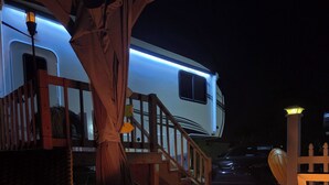 Een vuurkorf met een metalen grill, een rieten stoel, een houten tafel en een witte camper met blauwe verlichting.