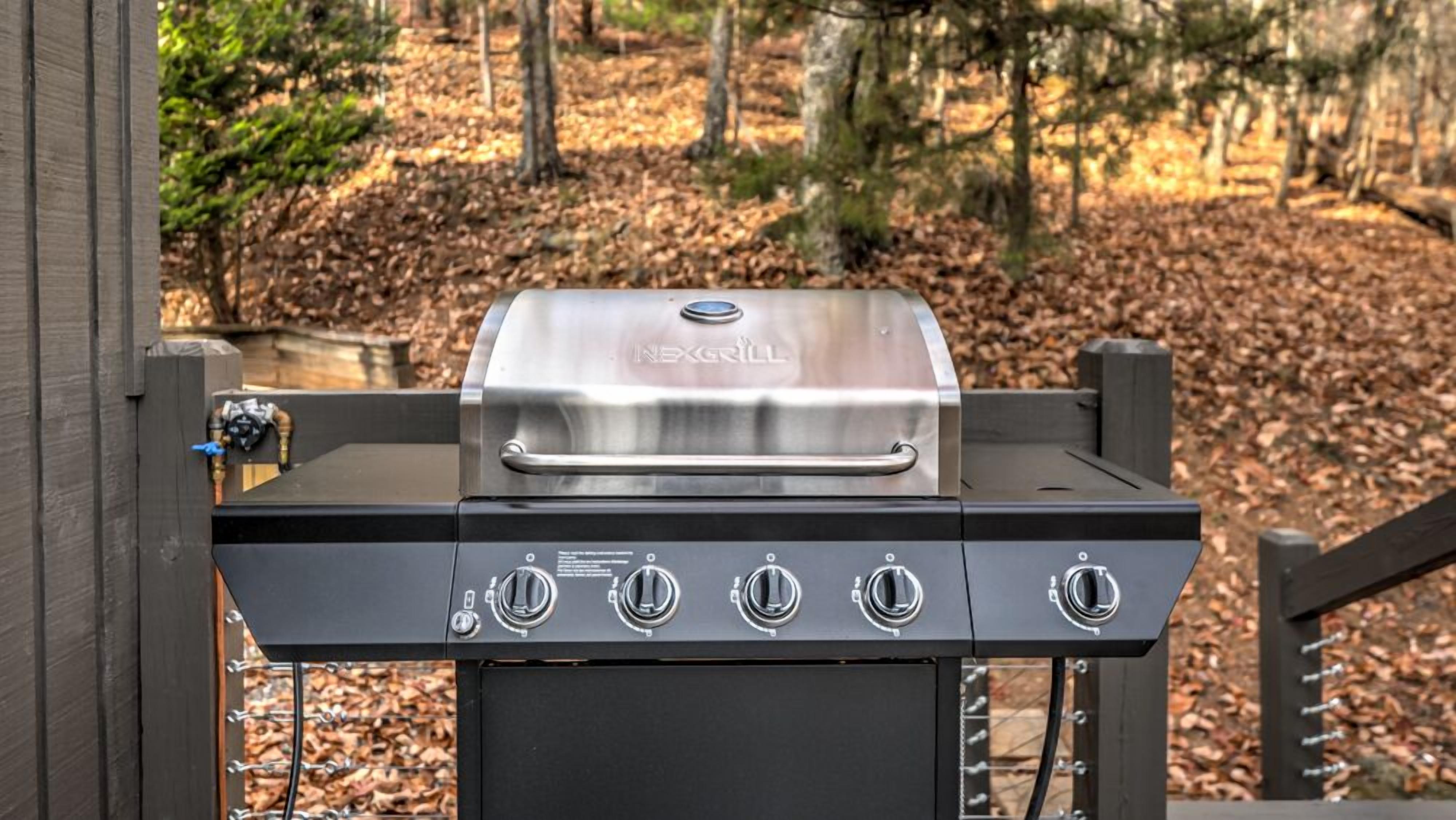 A NEXGRILL barbecue grill with four burners, a digital display, and a side burner, set against a wooden deck with a wooded background.