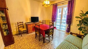 A room with a dining table, chairs, a television, a sofa, a plant, and a window with curtains.
