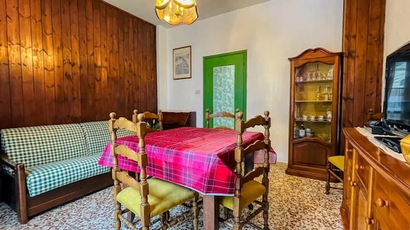 A room with a wooden wall, a checkered sofa, a table with a plaid tablecloth, a green door, and a wooden cabinet with glass doors.