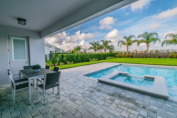 A swimming pool with a small hot tub, a patio with a table and chairs, and a view of a well-maintained lawn and palm trees.
