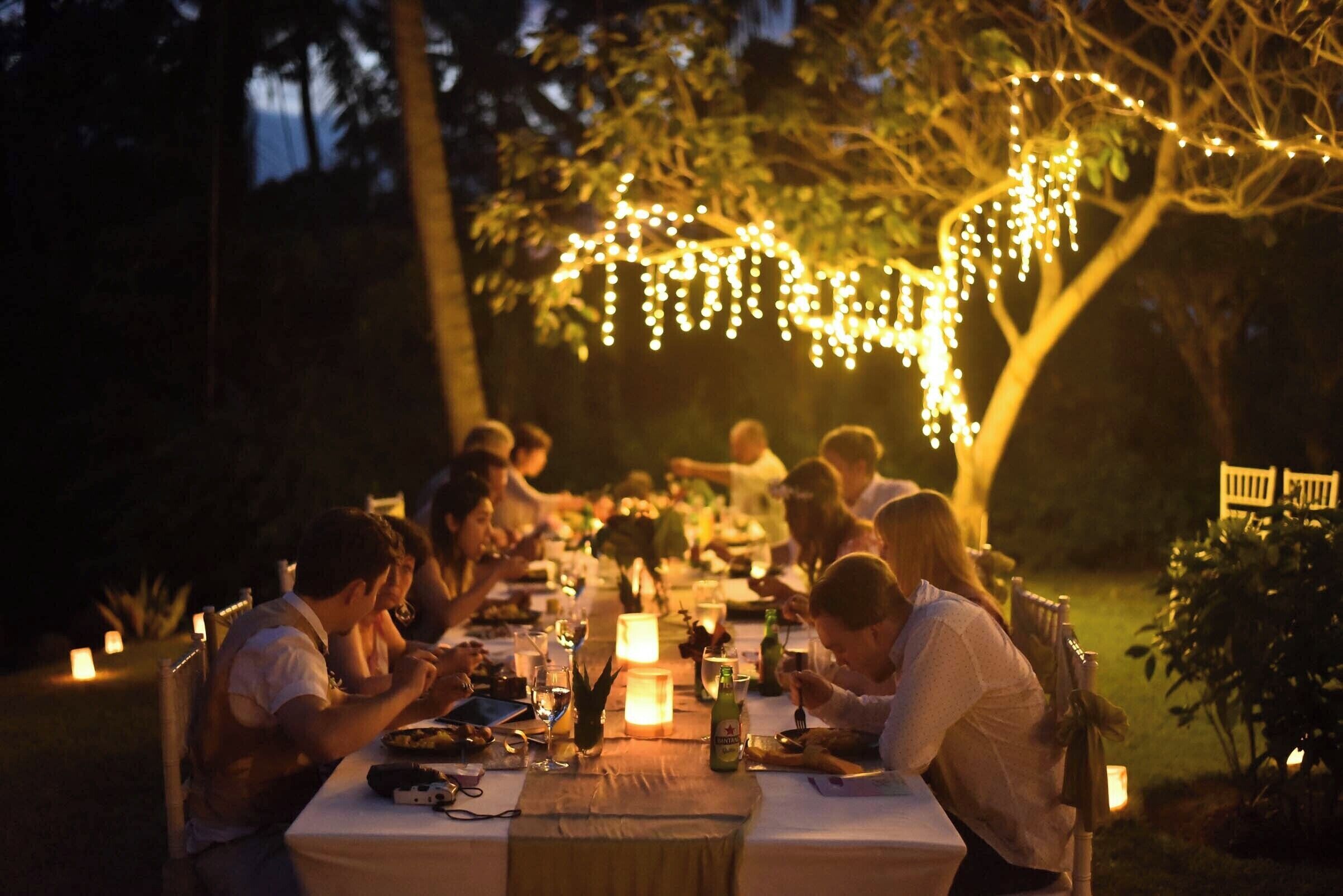 A group of people dining at an outdoor table with string lights overhead.
