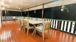 Un patio al aire libre con una mesa y sillas de madera, una cerca blanca de piquetes y plantas colgantes.