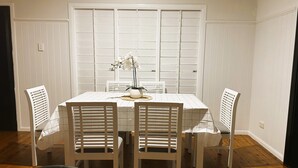Comedor con una mesa blanca y cuatro sillas blancas, una planta en maceta y una ventana con contraventanas.