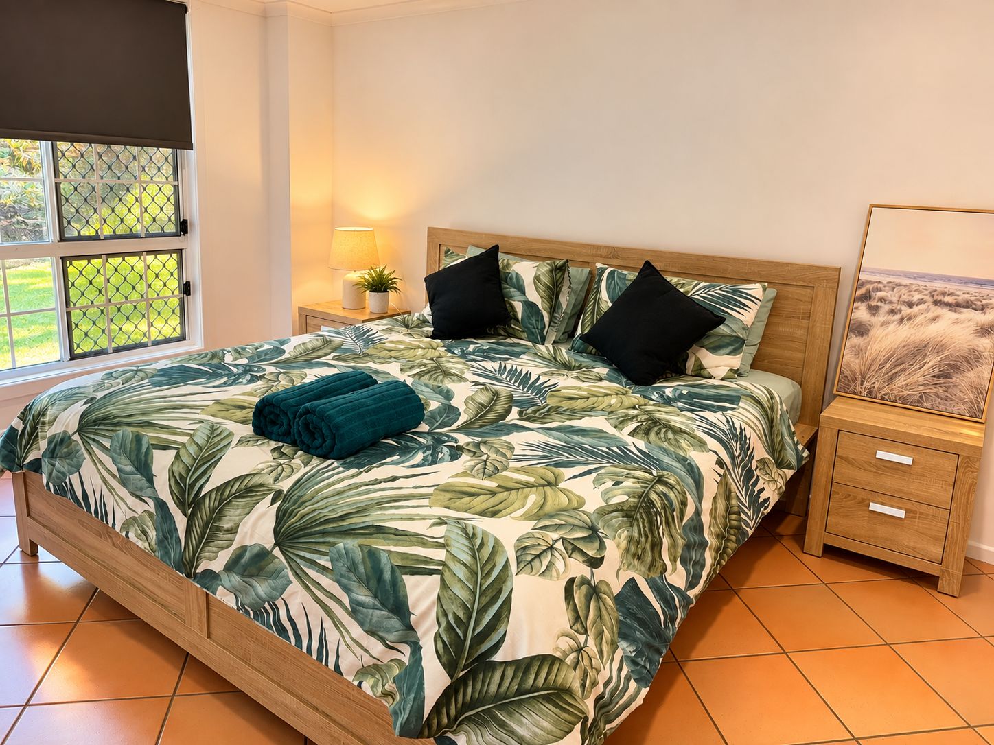 Un lit recouvert d’une couvre-lit ornée de motifs de feuilles tropicales, deux oreillers noirs, une serviette enroulée, une table de chevet avec une lampe et un tableau encadré accroché au mur.