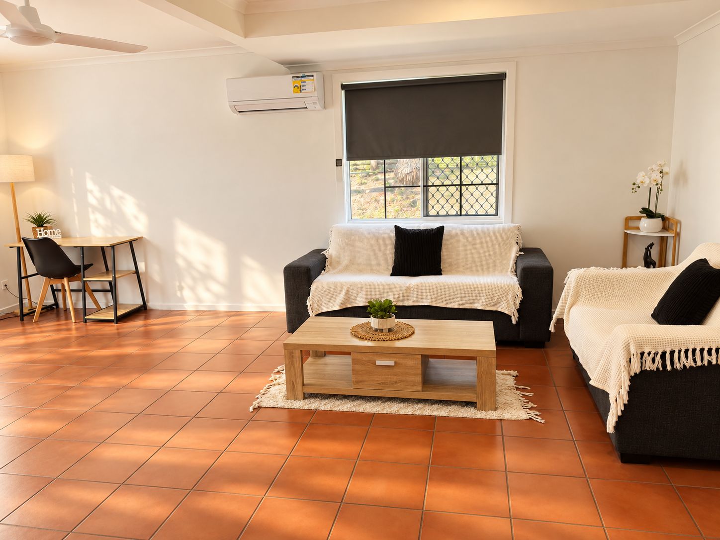 Un salon comprenant un canapé, une table basse et un fauteuil. On y trouve une fenêtre avec un store, un ventilateur de plafond et un appareil de climatisation.
