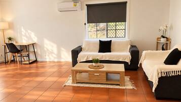 Un salon comprenant un canapé, une table basse et un fauteuil. On y trouve une fenêtre avec un store, un ventilateur de plafond et un appareil de climatisation.