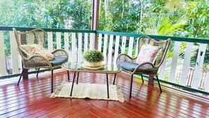 Una terraza de madera con dos sillas de mimbre y una mesa de vidrio, rodeada por una cerca blanca y abundante follaje.