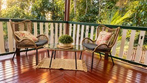 Une terrasse en bois, agrémentée de chaises en osier et d’une table en verre, entourée d’une clôture blanche et d’une végétation luxuriante.