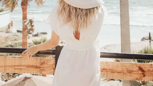 A person in a white dress and hat overlooking a beach with palm trees and the ocean. - Blvd Sunset. . . Beachfront Condos (Ensenada)