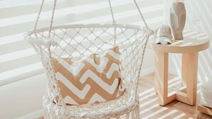 A macrame hanging chair with a chevron-patterned cushion, a small wooden side table, and a vase with dried flowers. - Blvd Sunset. . . Beachfront Condos (Ensenada)