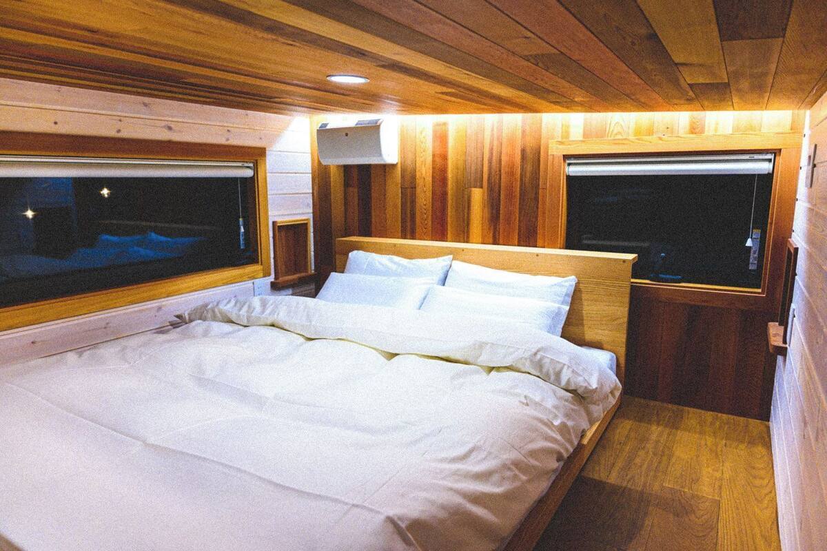 A wooden cabin interior with a bed, windows, and a ceiling light.