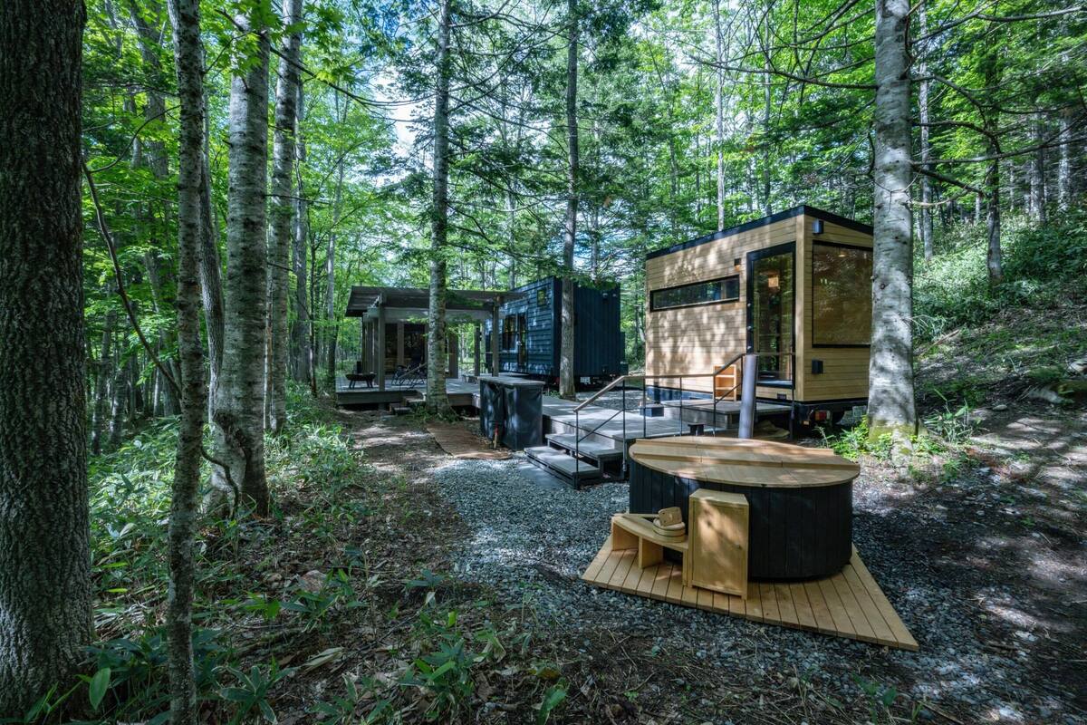 A forest cabin with a wooden deck, a hot tub, and a small outdoor seating area.