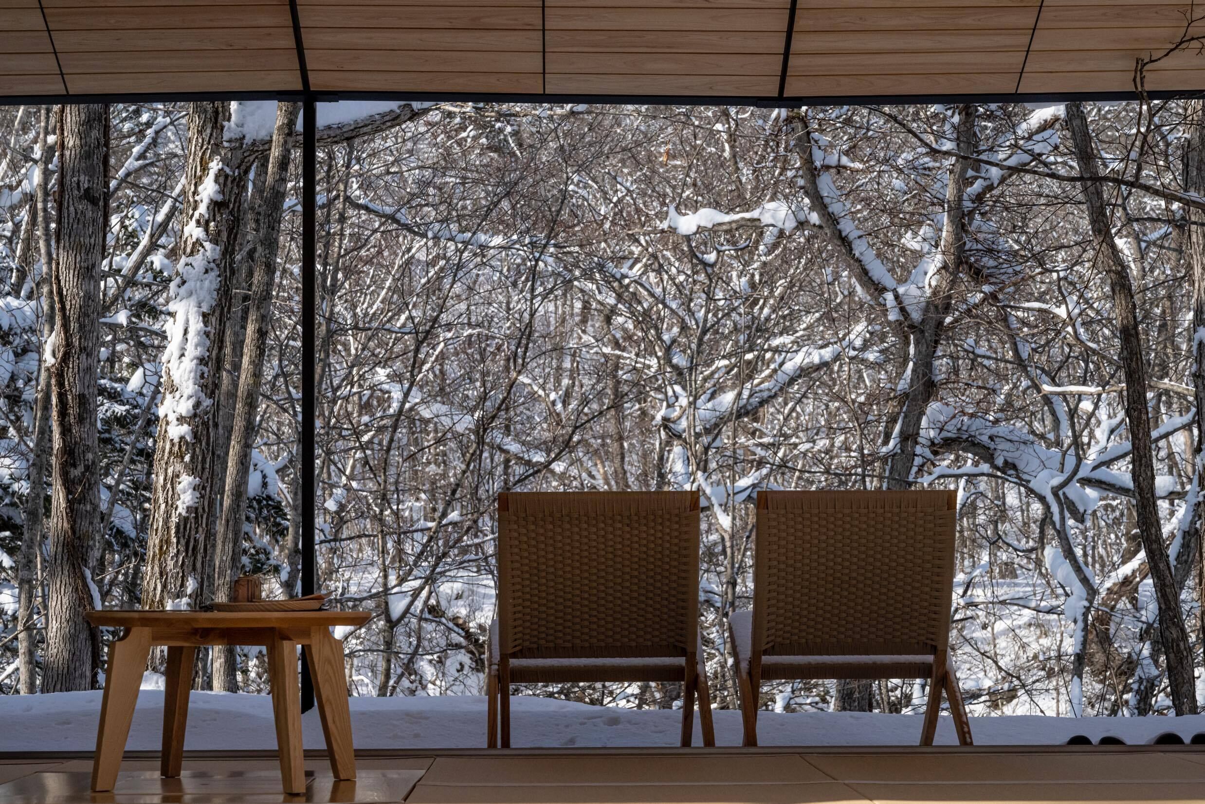 A wooden table and two wicker chairs in a covered outdoor area with a view of snow-covered trees.