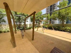 Zona de juegos al aire libre con columpios y una estructura para escalar, rodeada de vegetación y edificios.