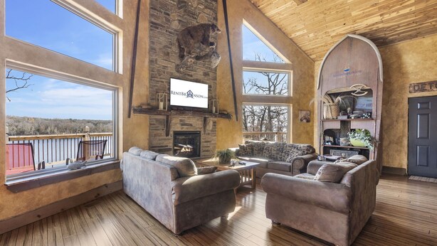 A spacious living room with a stone fireplace, large windows, and a view of the outdoors. - Lakefront Gem w/ Gorgeous Views, Boat Slip, Air Hockey & Close to Silver Dollar City (Branson)