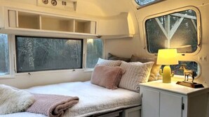 Ein gemütliches Wohnmobil-Interieur mit einem Bett, einem kleinen Tisch mit Lampe und einem Fenster mit Blick auf Bäume.