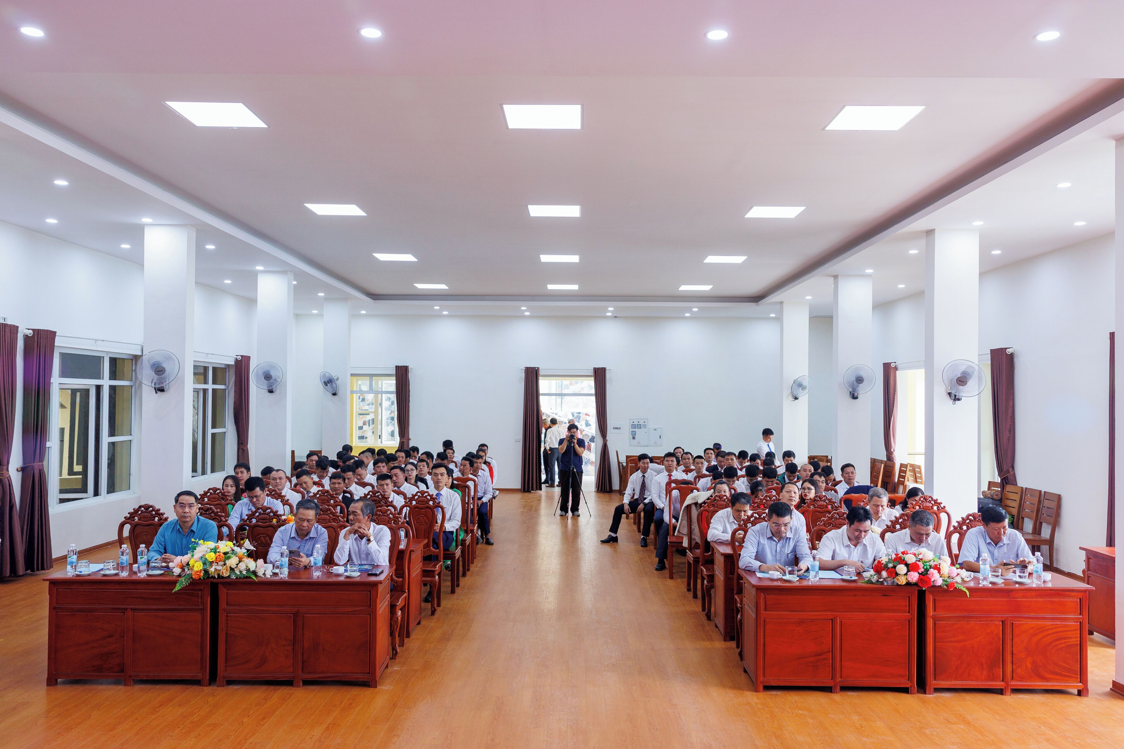 Un salón de conferencias con filas de mesas y sillas de madera, amplias ventanas y luces de techo.