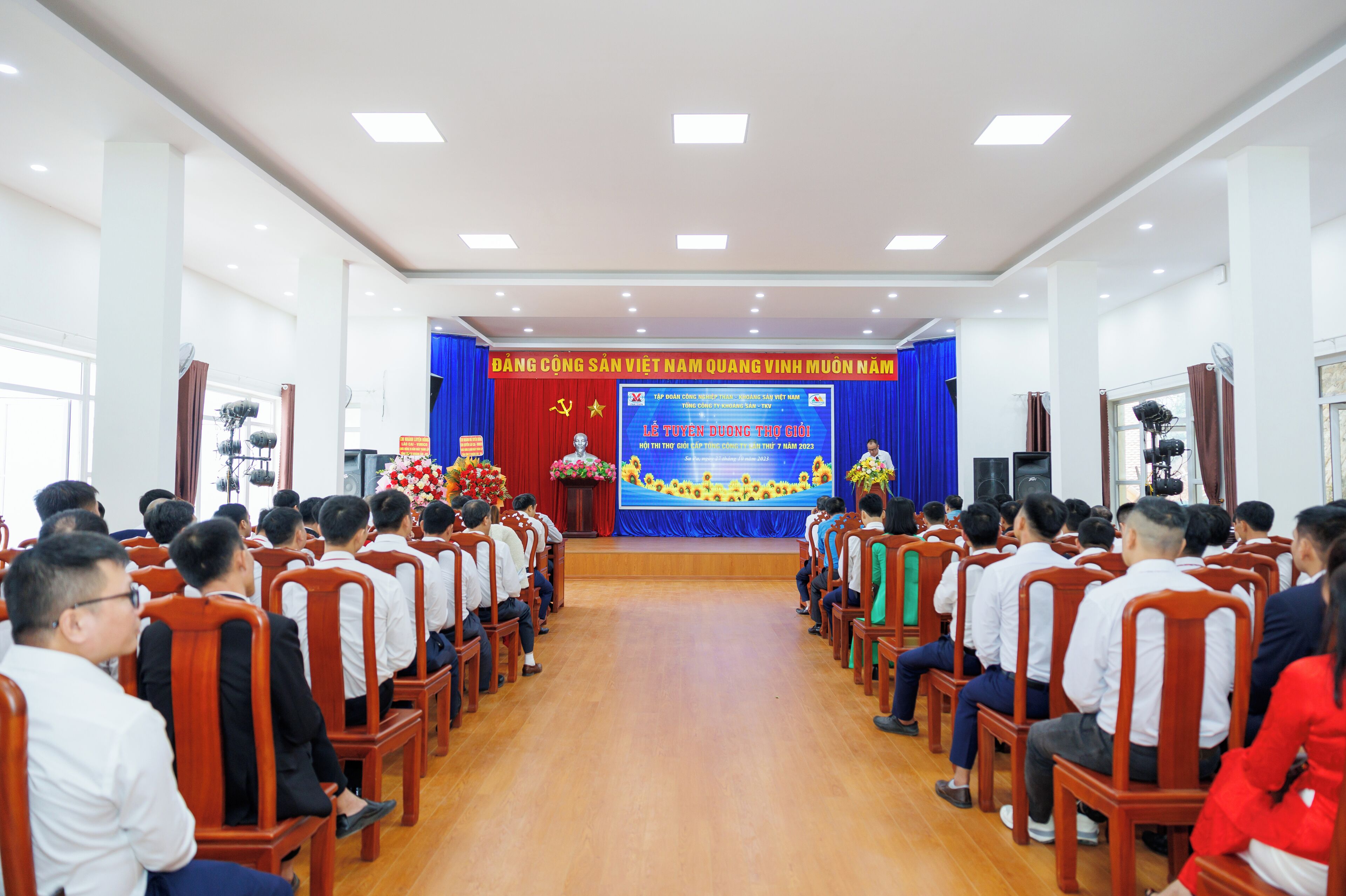A conference room with rows of wooden chairs facing a stage with a blue backdrop and a large screen.