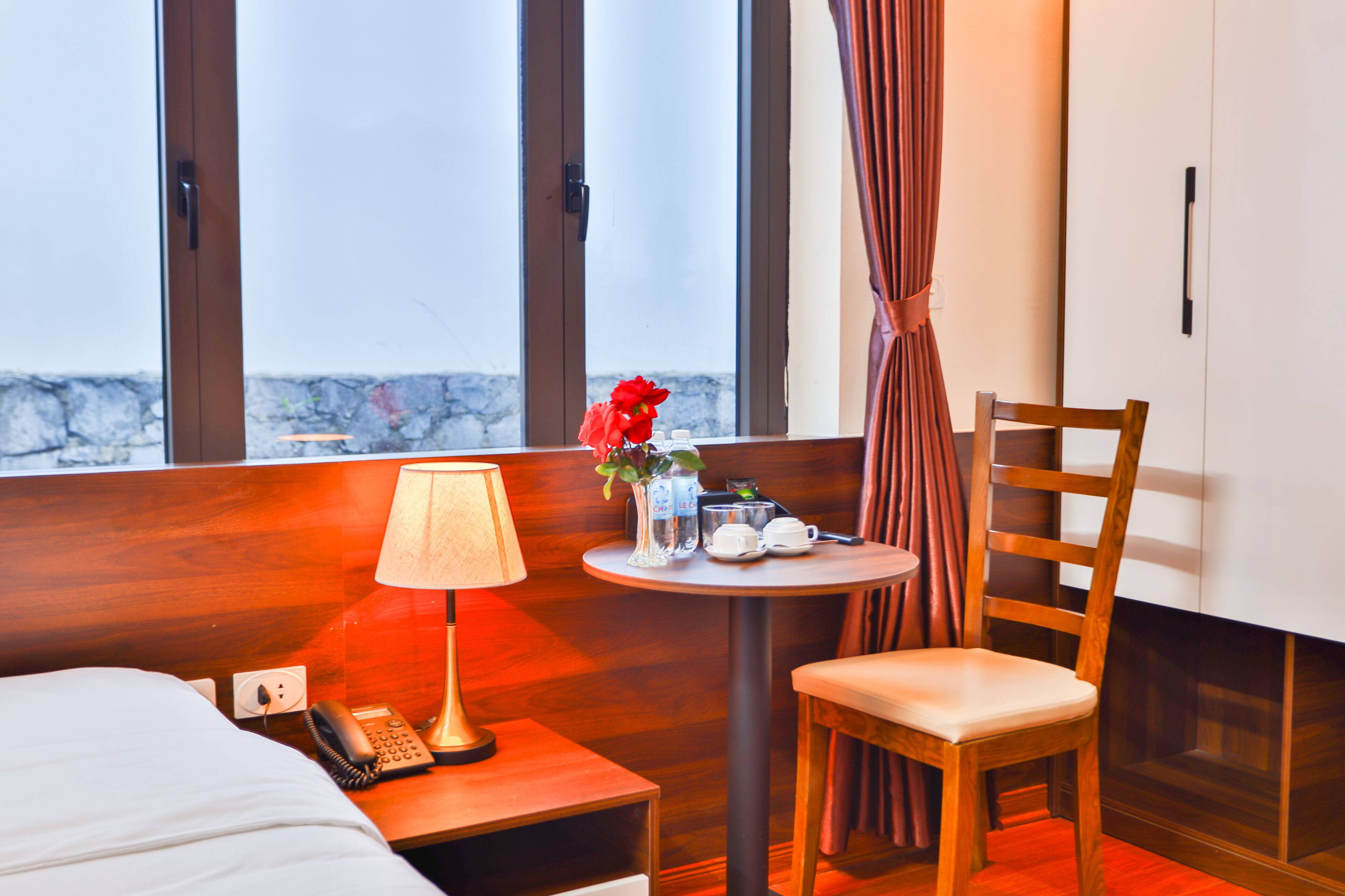 Une chambre d’hôtel avec un lit, une table de chevet avec une lampe, une petite table avec un vase de fleurs, une chaise et une fenêtre avec des rideaux.