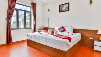 A neatly made bed with a white comforter and red accents, a bedside table with a lamp, and a window with curtains.