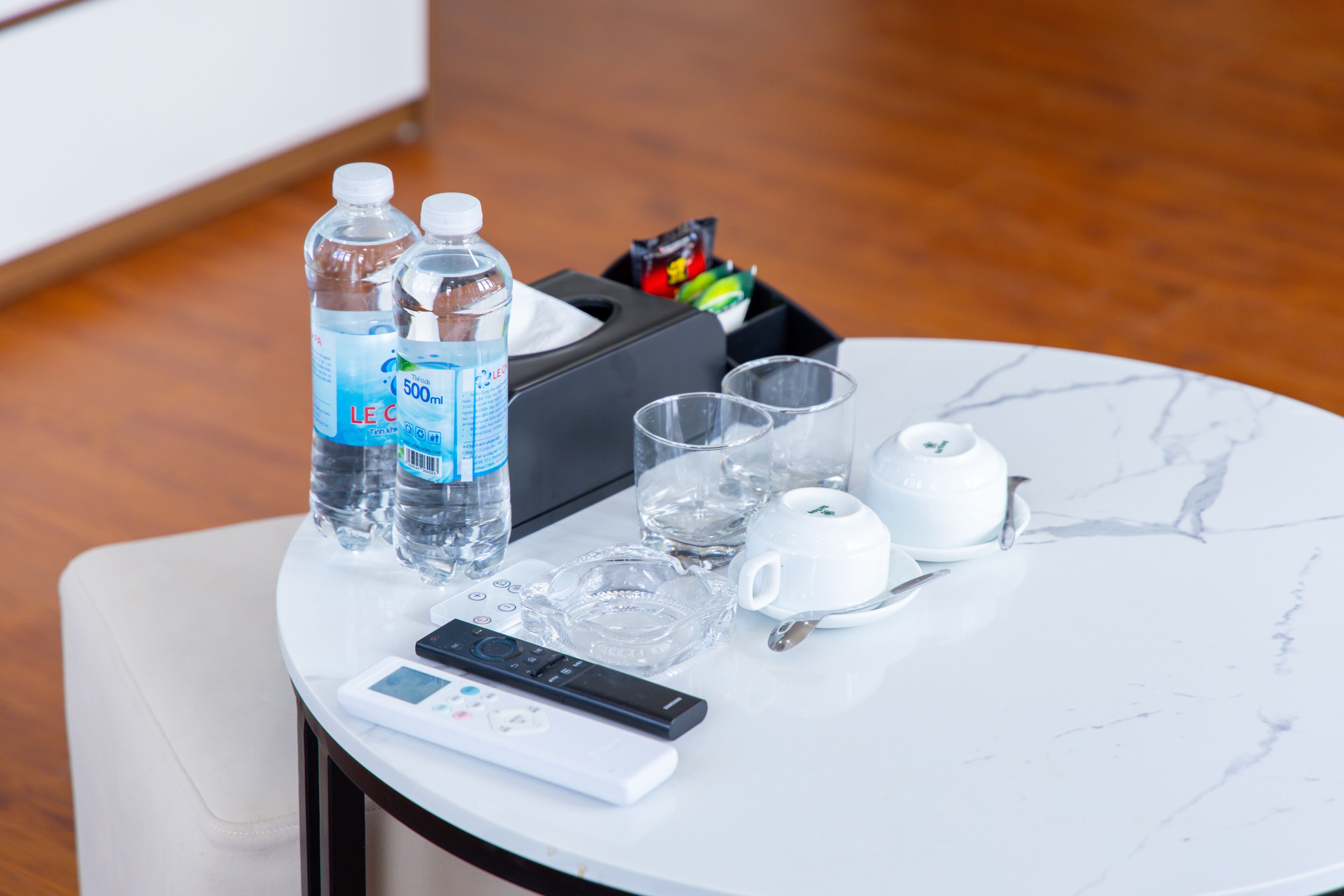 A table with two water bottles, a remote control, a glass, a teapot, and a cup on a marble table.