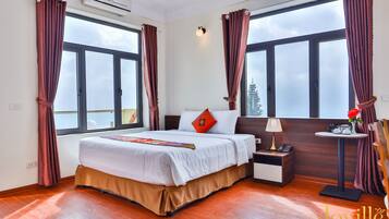 A hotel room with a large bed, a bedside table with a lamp, a small round table with a vase, and a view of the outdoors through large windows.