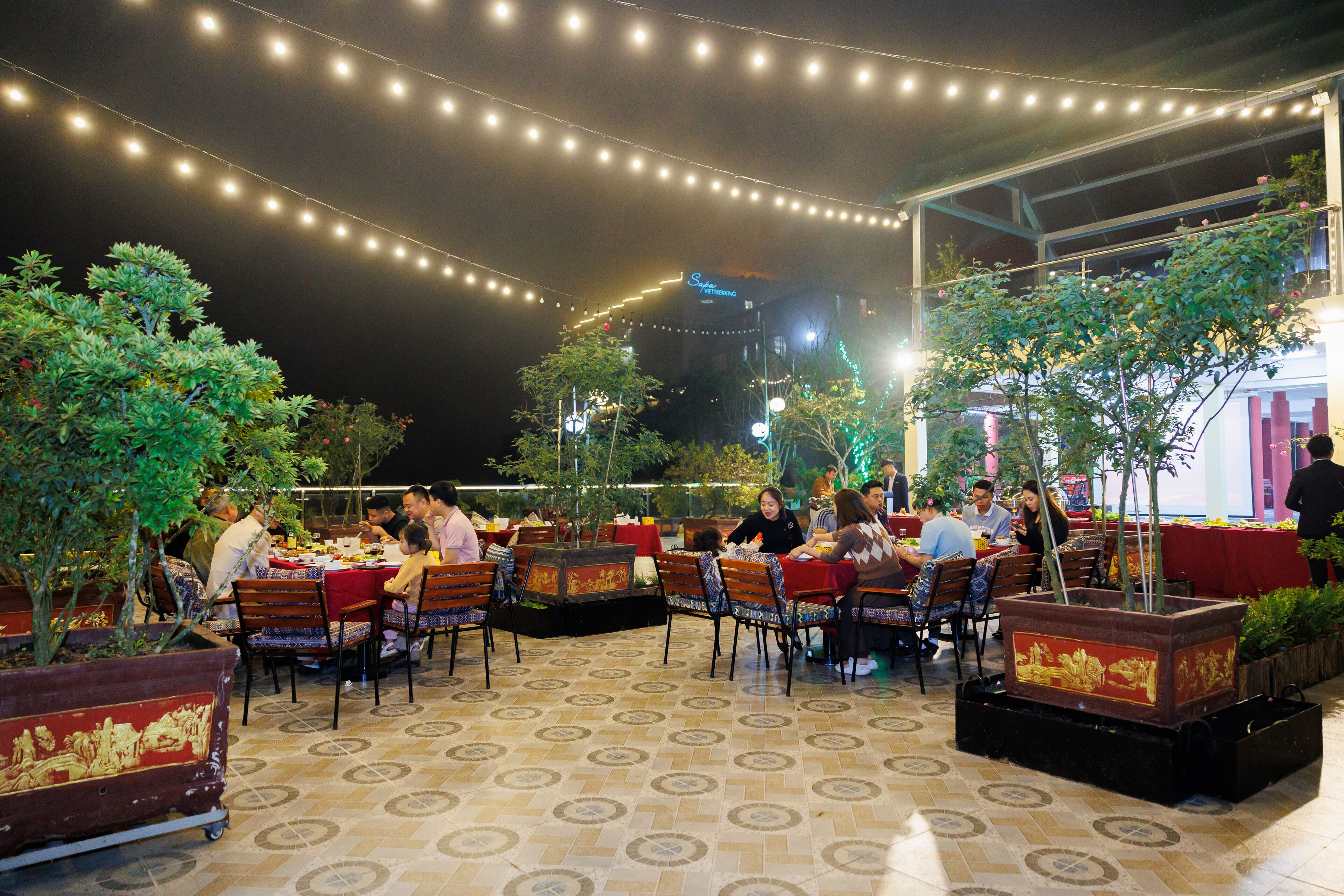 An outdoor dining area with tables and chairs, string lights overhead, and potted plants.