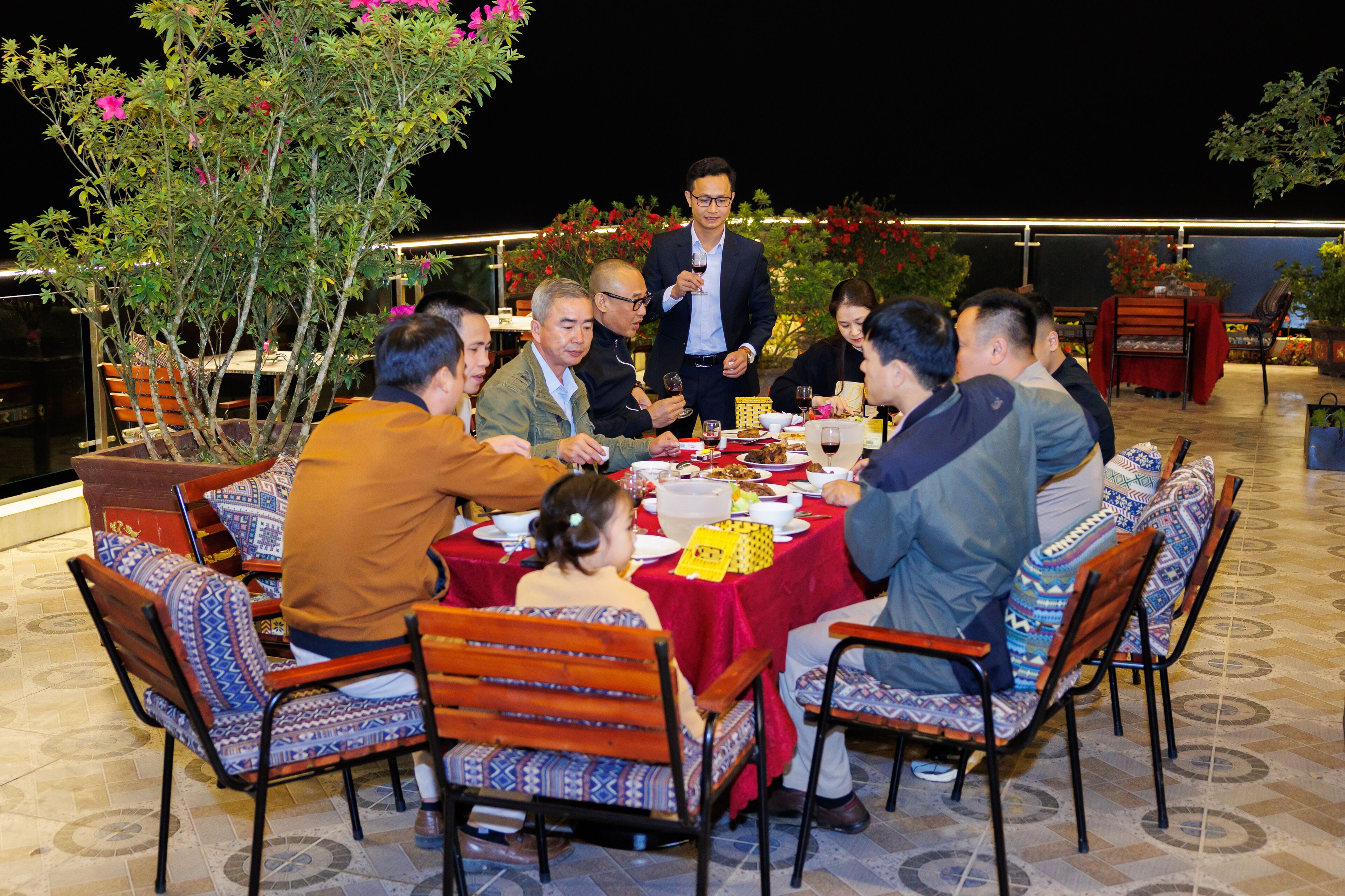 Un groupe de personnes est assis à une table à l’extérieur, recouverte d’une nappe rouge, et des chaises ornées de coussins à motifs les entourent.