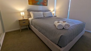 A neatly made bed with a checkered bedspread, two pillows, and rolled towels on the side. There is a nightstand with a lamp and a drawer. A colorful abstract painting hangs above the bed. - Newcastle Interchange Apartments (Wickham)