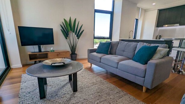 A modern living room with a grey sofa, a round coffee table, a flat-screen TV, and a large window. - Newcastle Interchange Apartments (Wickham)