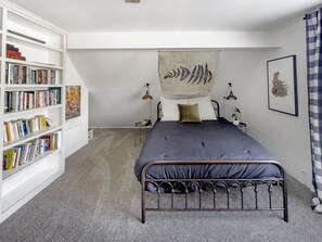 A bedroom with a metal bed frame, a bookshelf filled with books, a wall-mounted fern artwork, and a framed picture on the wall.