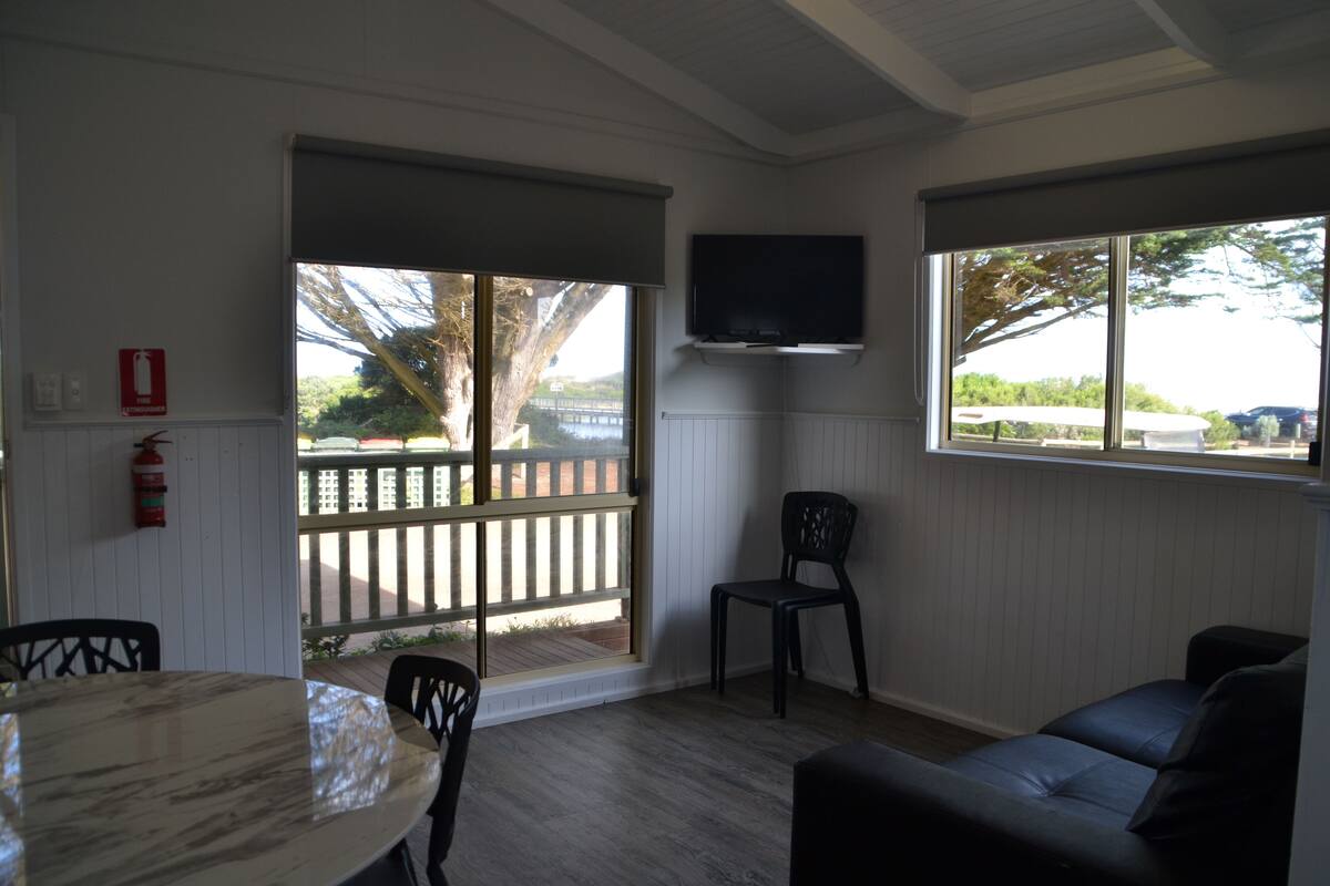 Una habitación con piso de madera, una mesa redonda, una silla negra, un sofá negro, un televisor de pantalla plana montado en la pared y una ventana con vista a una terraza y árboles.