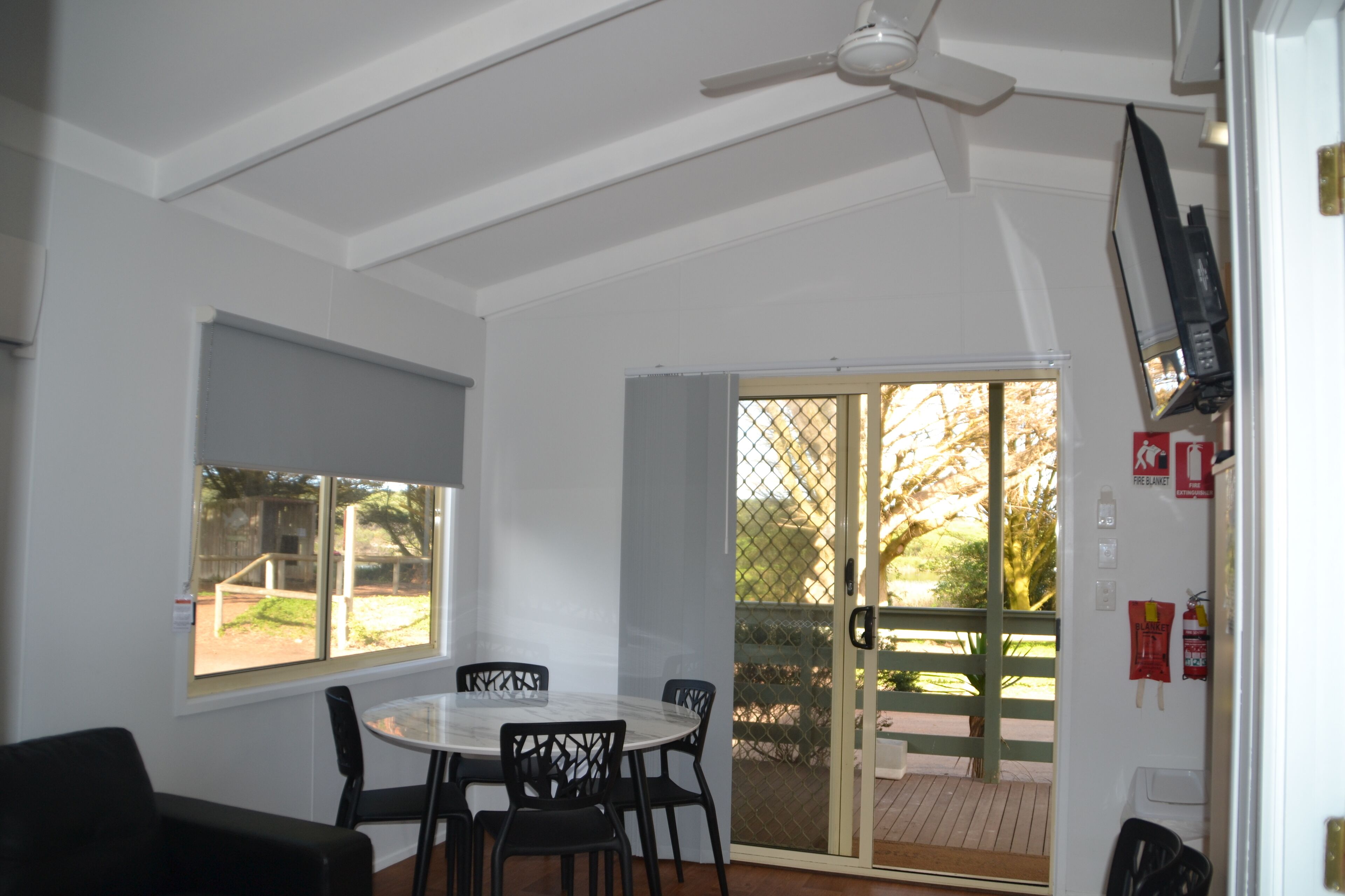A room with a white ceiling, a ceiling fan, a table with chairs, a sliding glass door leading to an outdoor area, and a fire extinguisher mounted on the wall.
