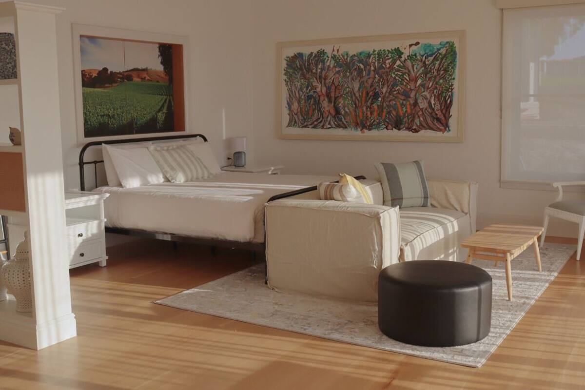 A modern bedroom with a bed, a sofa, a small table, and a chair. There is a painting on the wall and a window with a view.