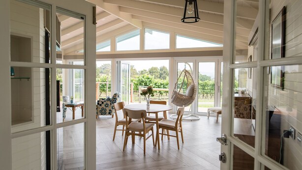 Un espacio de sala comedor amplio con una mesa, sillas, un sillón colgante y vistas a un viñedo a través de amplios ventanales.