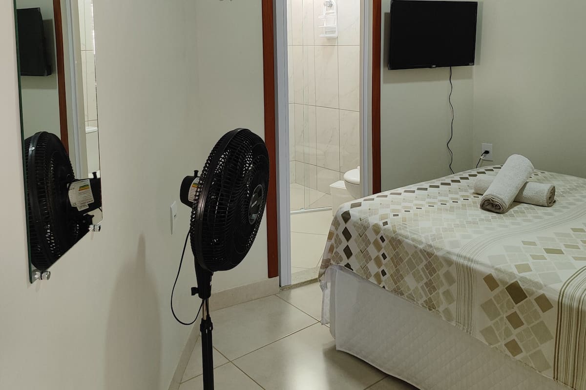 Une chambre à coucher comprenant un lit, un ventilateur sur pied, un téléviseur fixé au mur et une porte donnant sur une salle de bain.
