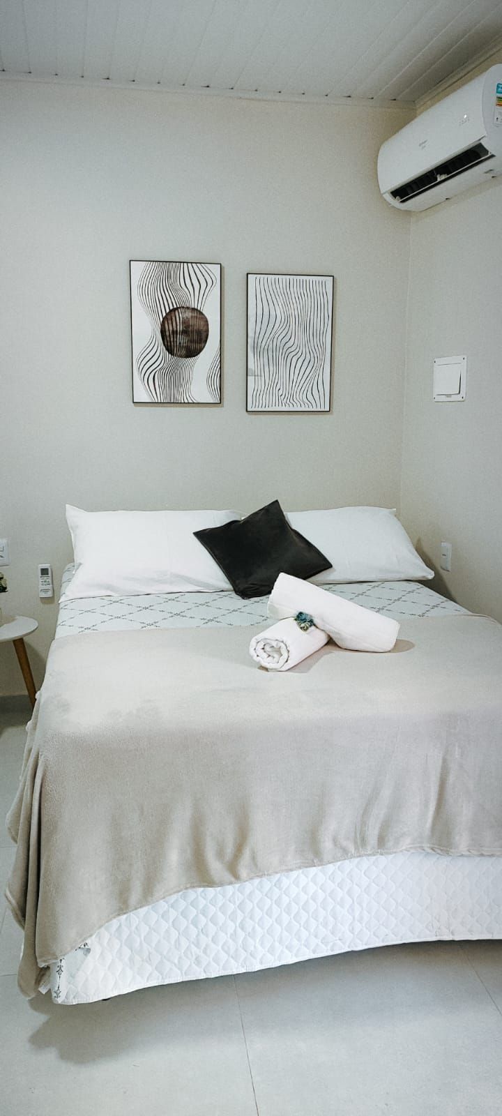 Ein ordentlich bezogenes Bett mit einer gesteppten Tagesdecke, zwei Kissen und einem zusammengerollten Handtuch. Darüber hängen zwei an der Wand befestigte Kunstwerke.
