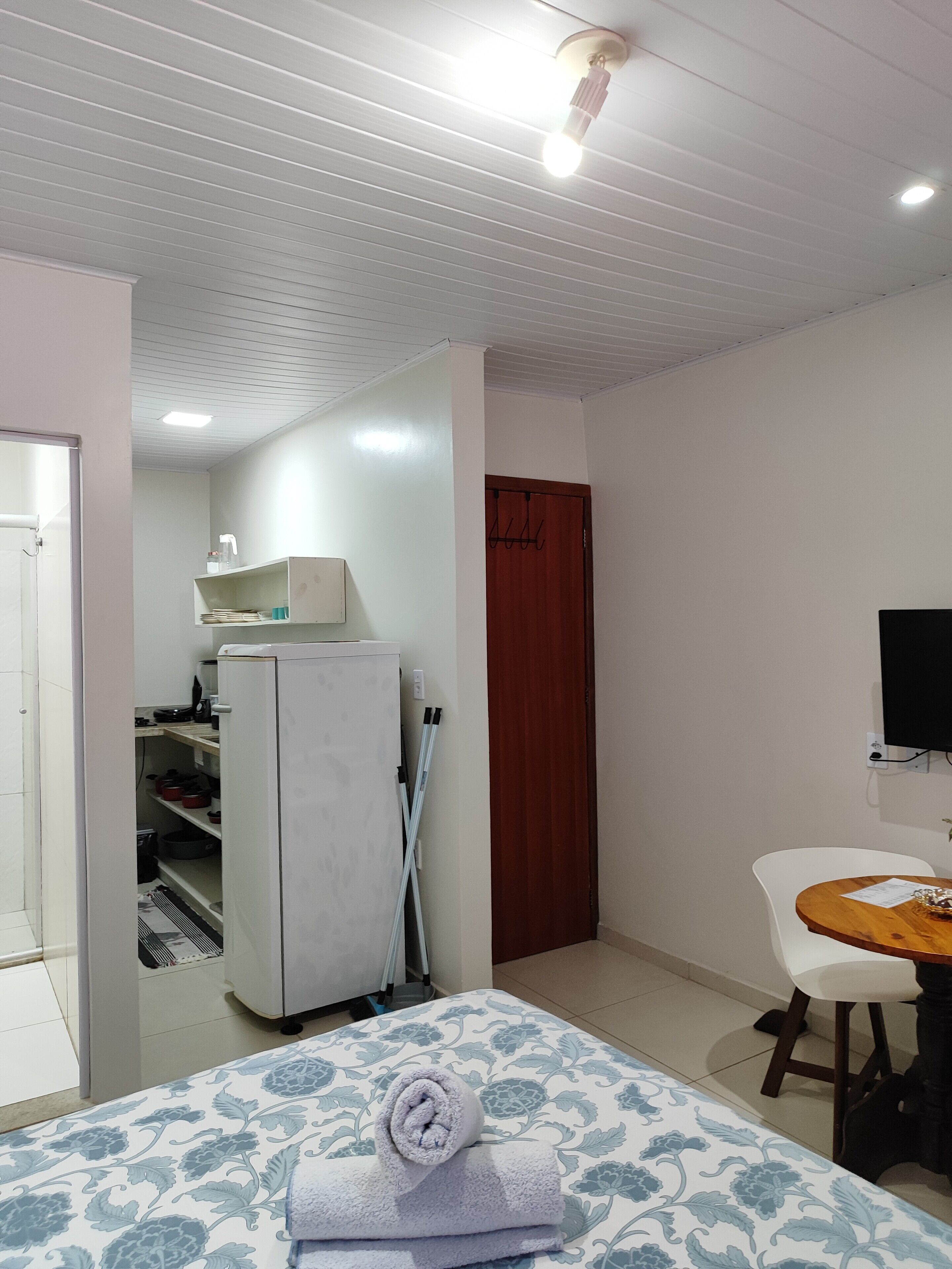 A room with a bed, a small table with a chair, a television, a refrigerator, and a door leading to a kitchen area.