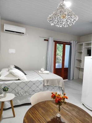 A hotel room with a bed, a small table with a vase of flowers, a refrigerator, and a window with curtains. - Casa de Mel (Itacaré)