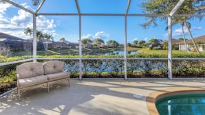 Ein überdachter Außenbereich mit Pool, einem beigen Sofa und Blick auf einen See sowie Häuser im Hintergrund.