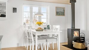 A dining area with a white table and chairs, a vase with flowers, a black stove, and a window with a view of trees. - Gorgeous home in Karrebæksminde (Karrebæksminde)