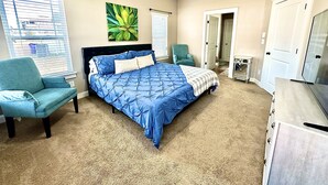 Een slaapkamer met een bed, twee stoelen, een bureau, een televisie en een deur naar een andere kamer.