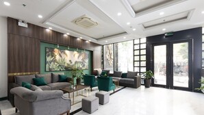 Un vestíbulo moderno con piso de mármol, sofás grises, sillones verdes y un cuadro grande de plantas tropicales.