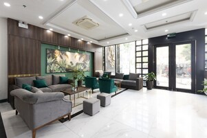 Un vestíbulo moderno con piso de mármol, sofás grises, sillones verdes y un cuadro grande de plantas tropicales.