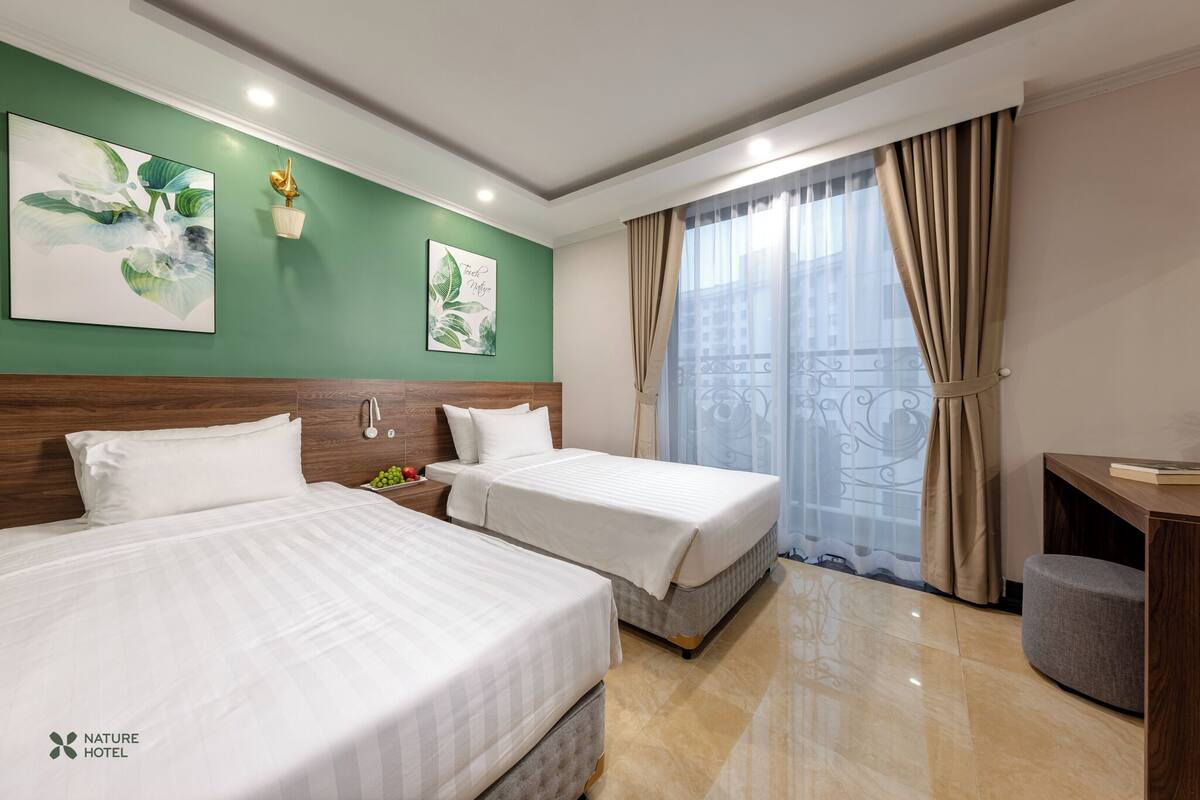 A hotel room with two beds, a green accent wall, a window with curtains, and a small desk with a chair.