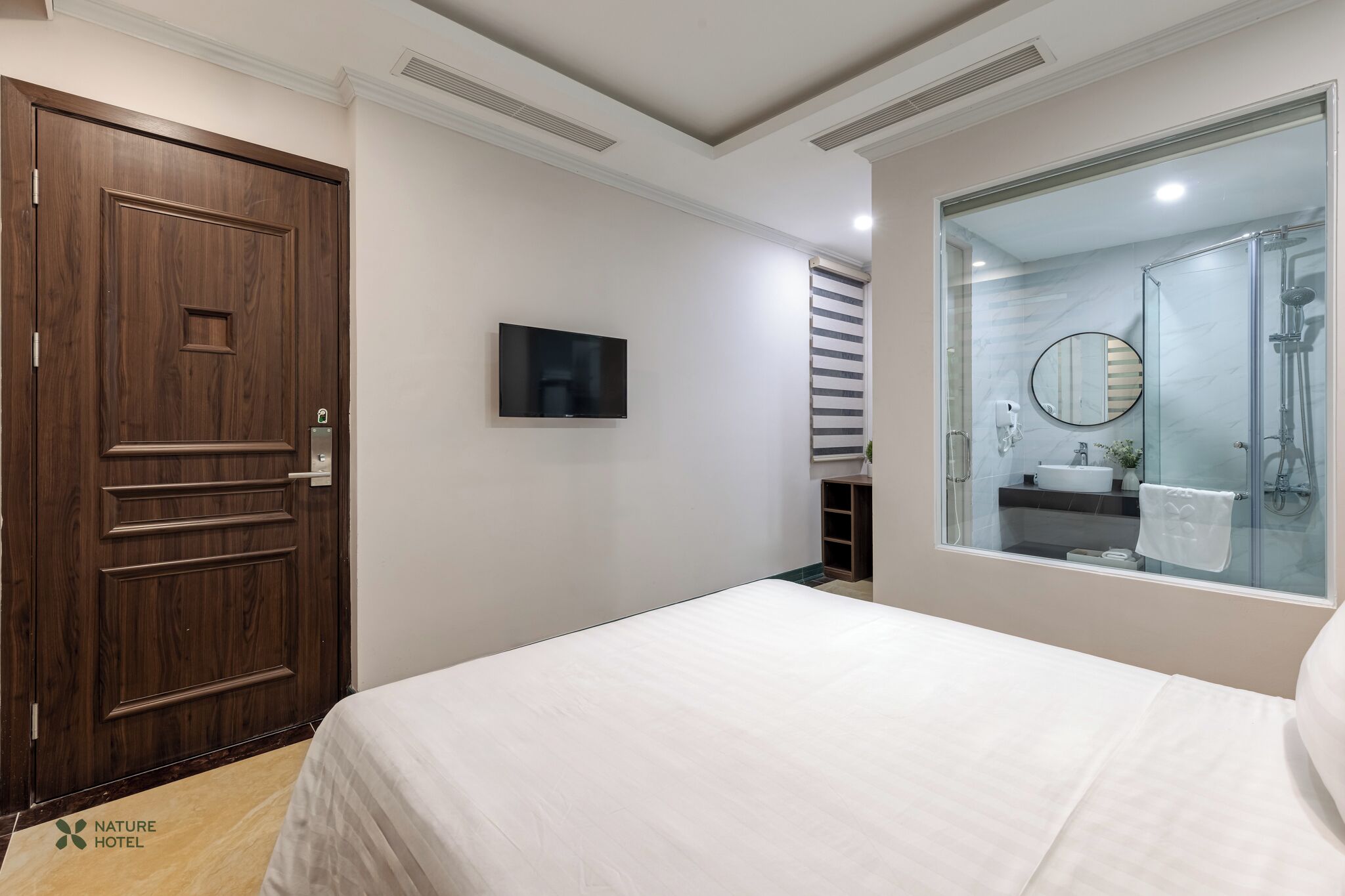 A hotel room with a large bed, a wooden door, a television, and a bathroom with a glass shower enclosure.