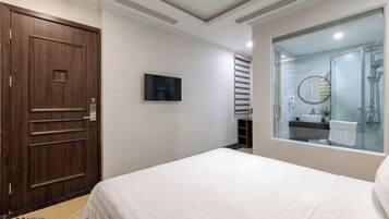 A hotel room with a large bed, a wooden door, a television, and a bathroom with a glass shower enclosure.
